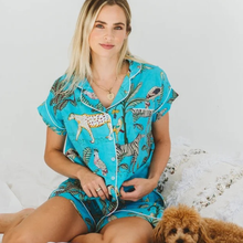Load image into Gallery viewer, A woman wearing a turquoise blue short-sleeve pajama set with a safari animal print, sitting on a bed with a small brown dog next to her.