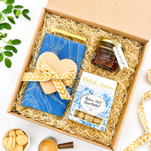 Load image into Gallery viewer, A curated gift box set in a brown cardboard box filled with kraft paper shred, featuring a blue &amp; white A Love Supreme tea towel with a wooden heart-shaped coaster and yellow ribbon, a jar of Delish Sisters chilli crunch, a box of Delish Sisters pecan nut shortbread, and gold heart-shaped tongs on a white background.
