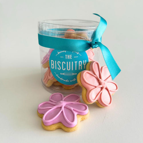 Clear container with cookies decorated like flowers, tied with a blue ribbon, branded 'The Biscuitry'.