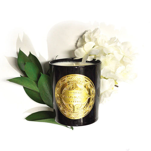 Black candle with gold label surrounded by white flowers and green leaves on a white background