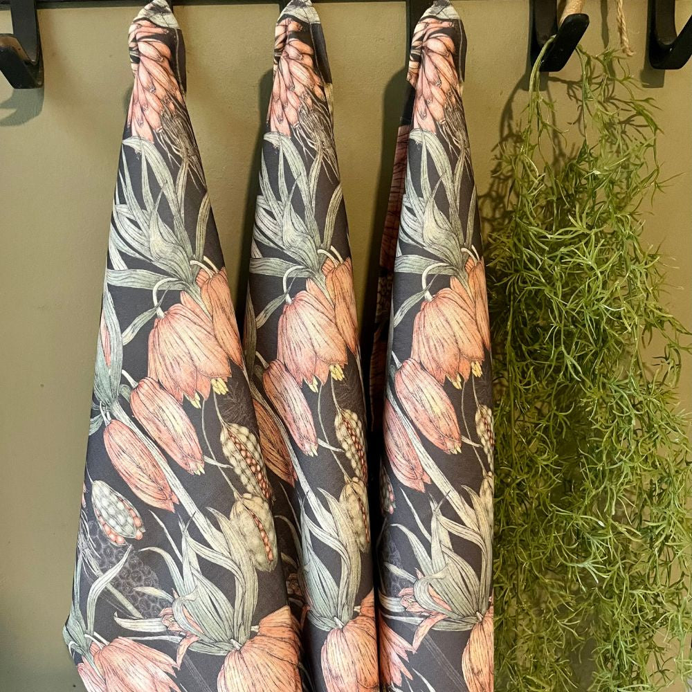 Three JW Designs Grey Aloe tea towels hanging on a rack with greenery in the background.