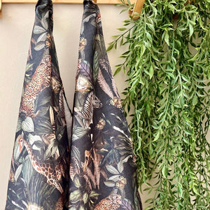 JW Designs black African safari print tea towels hanging on a wooden rod with a green plant in the background.