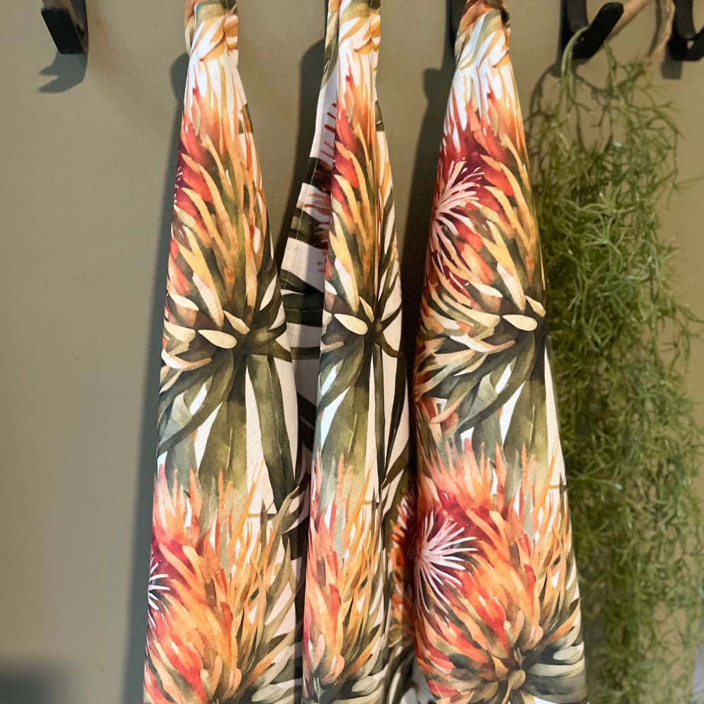 Three JW Designs Orange Pincushion print tea towels hanging on a rack with a plant in the background.
