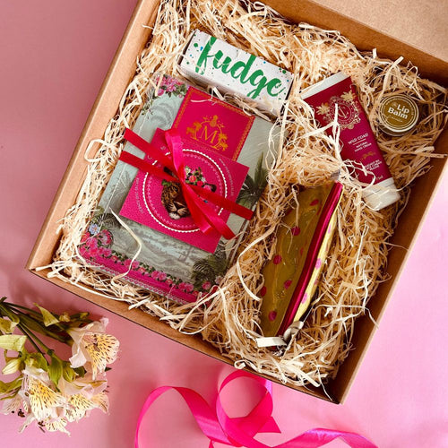 Gift box with various items including a book, fudge, hand cream, lip balm and a make up bag on a pink background