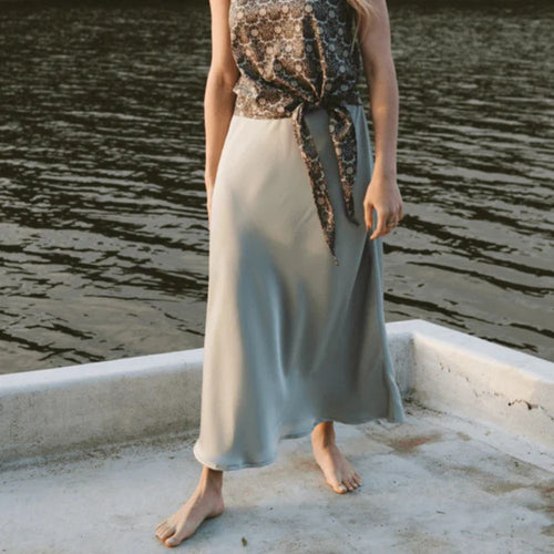 Person wearing a long, mint satin skirt with a patterned top by a body of water.