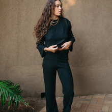 Load image into Gallery viewer, Woman wearing a dark outfit standing against a beige wall with a plant to her left.
