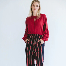 Load image into Gallery viewer, Woman wearing a red blouse and black striped pants against a light blue background
