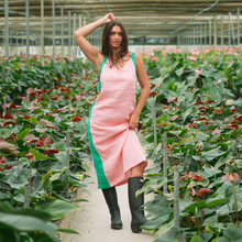 Load image into Gallery viewer, Woman in a pink dress with green accents standing in a greenhouse filled with plants.
