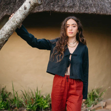 Load image into Gallery viewer, Woman in a black jacket and red pants standing under a thatched roof.
