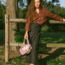 Load image into Gallery viewer, Woman in a brown shirt and black wrap skirt holding a basket with eggs, leaning against a wooden fence in a natural setting.
