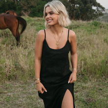 Load image into Gallery viewer, Woman in a black dress standing in a field with a horse in the background
