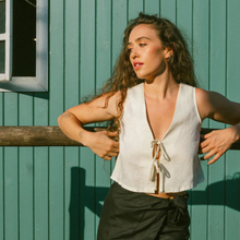 Load image into Gallery viewer, Woman in a white sleeveless top and black skirt standing against a teal wooden wall.
