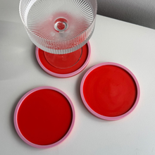 Load image into Gallery viewer, Red coasters with pink rims on a white surface, one with a glass.
