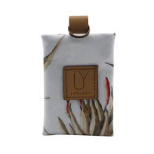 Load image into Gallery viewer, A small fabric pouch with a light gray background and a botanical print in beige, brown, and muted red. It features a tan leather patch on the front with the brand name 'IY APPAREL' and a matching tan loop at the top.
