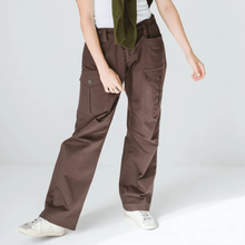 Load image into Gallery viewer, A person wearing brown wide-leg cargo pants with side flap pockets, styled with a white top, an olive green shirt draped over the shoulder, and white sneakers against a light gray studio background.
