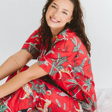 Load image into Gallery viewer, A woman wearing a red short-sleeve button-up pyjama set with a colourful jungle and animal print, sitting against a plain white background.
