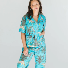Load image into Gallery viewer, A woman modeling a turquoise short-sleeve pajama set with an all-over safari print featuring leopards, toucans, and palm trees against a light gray studio background.
