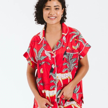Load image into Gallery viewer, A woman wearing a red short-sleeve pajama shirt with a tropical print of toucans, birds, and jaguars, featuring white piping on the collar, buttons, and pocket, set against a plain white background.
