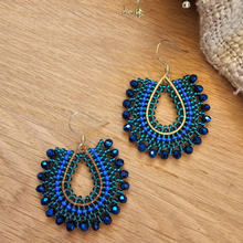 Load image into Gallery viewer, A pair of handmade teardrop-shaped dangle earrings with blue and green seed beads, dark blue faceted beads, and gold-tone metal wire frames, displayed on a wooden surface.
