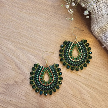 Load image into Gallery viewer, A pair of handmade beaded dangle earrings with green faceted beads and gold-tone metal, displayed on a light wood surface next to small white flowers and a woven fabric corner.
