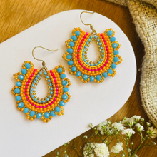 Load image into Gallery viewer, A pair of handmade teardrop-shaped beaded earrings with turquoise, pink, orange, and gold beads, displayed on a white curved plate over a wooden surface with burlap and baby's breath flowers.
