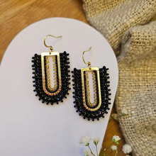 Load image into Gallery viewer, A pair of handmade beaded dangle earrings with gold-tone hooks and frames, featuring black seed beads and gold-toned metallic accents, displayed on a white display card with a burlap fabric background.
