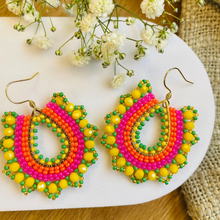 Load image into Gallery viewer, A pair of handmade beaded hoop earrings with gold-tone fishhook wires, featuring concentric bands of yellow, orange, pink, and green beads. The earrings are displayed on a white ceramic plate with baby's breath flowers and a burlap fabric background.
