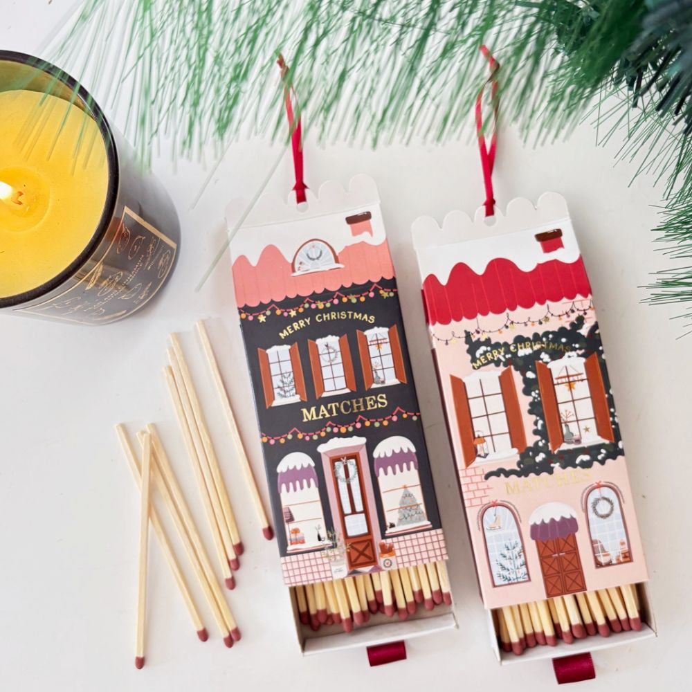 Two Christmas-themed matchboxes with matches on a white surface with greenery.